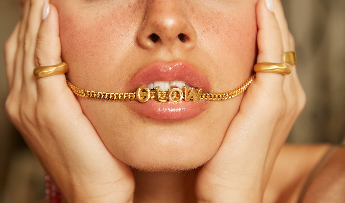 A close-up of a model with a gold chain featuring the word 'Glow' in her mouth, showcasing elegant jewelry by CINCO.