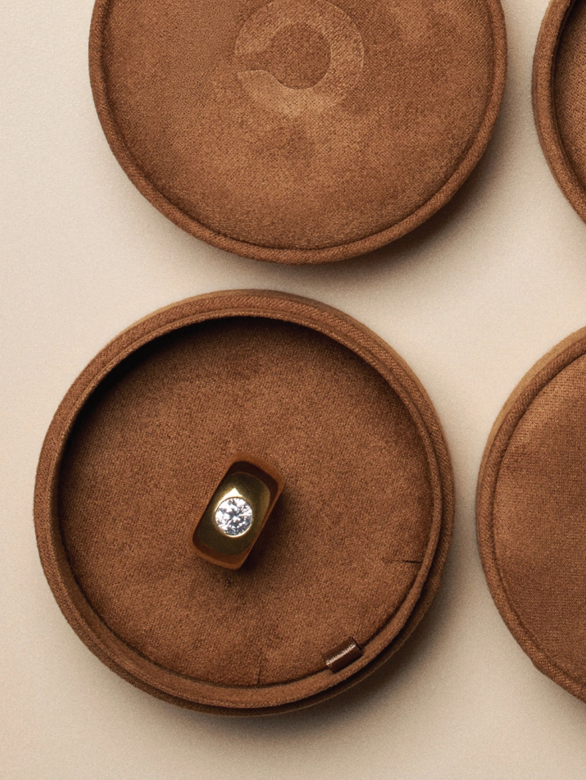 A gold ring with a clear round gemstone displayed inside an open round brown velvet ring box, with additional closed matching boxes on a beige background.