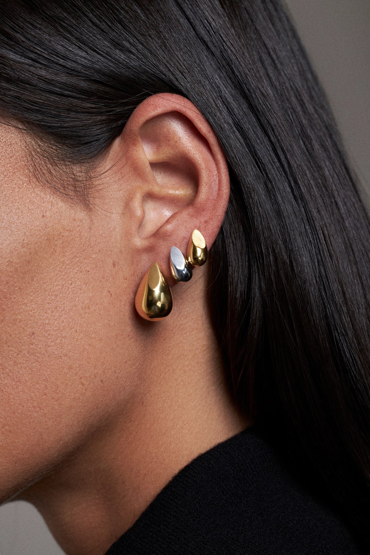 A close-up of a person's ear wearing three teardrop-shaped metallic earrings in gold, silver, and black, with long dark hair and a black top visible.