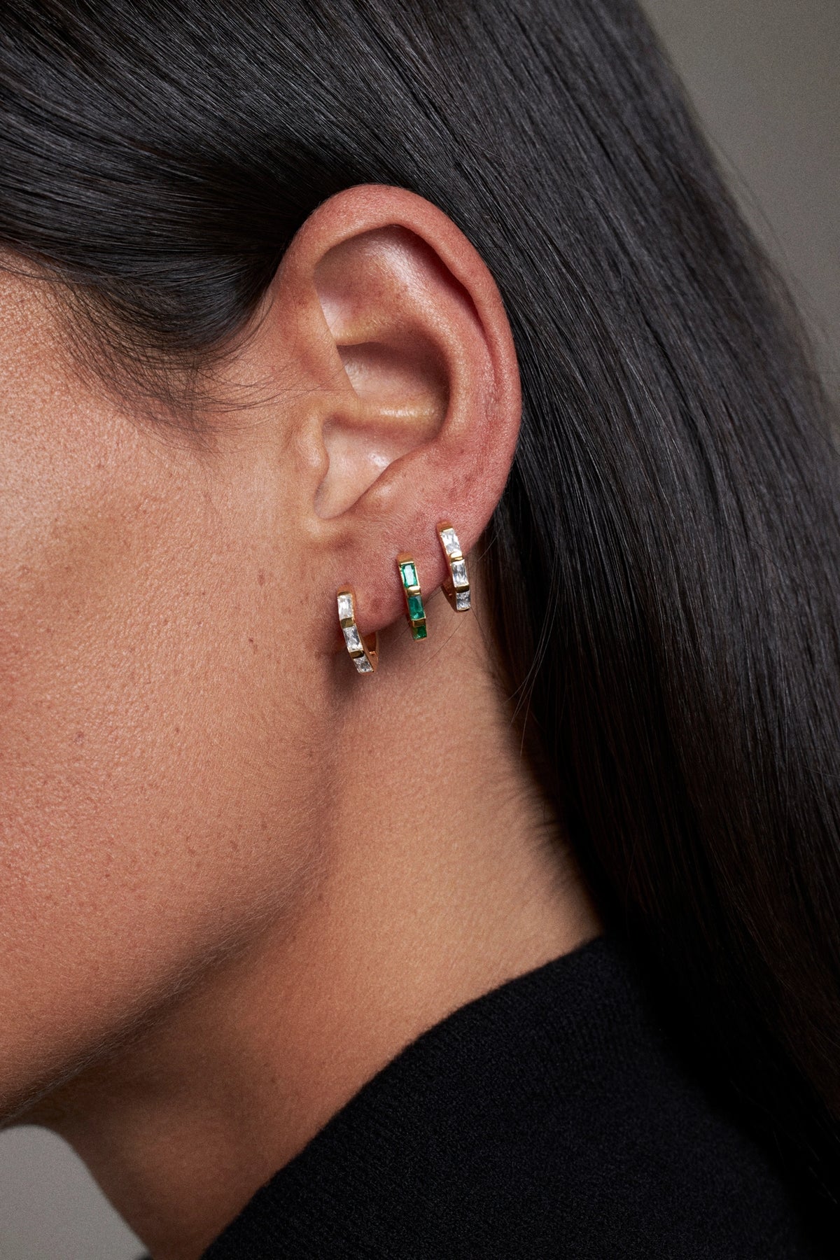 Close-up of a model's ear wearing three small hoop earrings with clear stones and green rectangular accents, against dark hair and a black top.