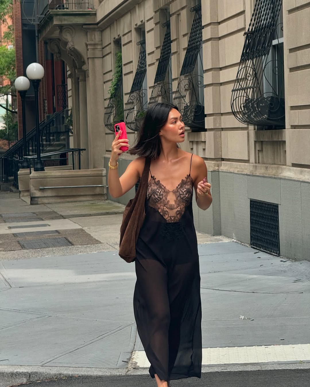 Woman wearing a sheer black outfit with lace detailing, walking down a city street, holding a phone.