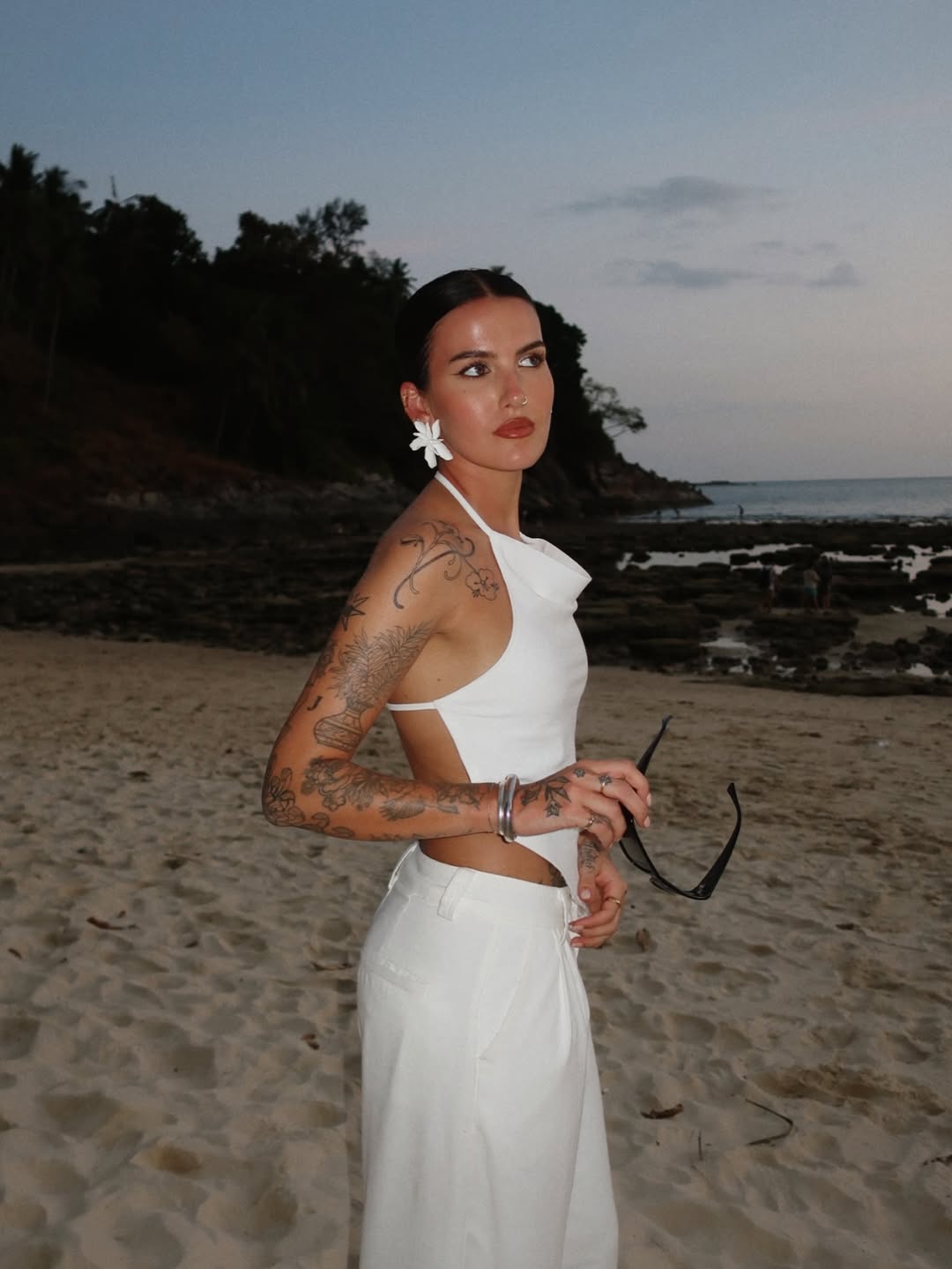 Woman in white outfit standing on beach at dusk, holding sunglasses and adorned with tattoos and flower in hair.